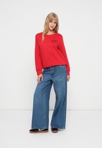 Červená mikina s černým logem "Gap", v kombinaci s volnými modrými džíny. Modelka stojí na jednobarevném pozadí a má na sobě černé slip-on boty.