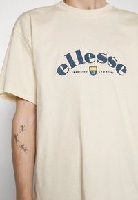 Camiseta de algodón color crema con mangas cortas, que presenta un gran logo en forma curva en color marino y un pequeño gráfico de un escudo en el centro.