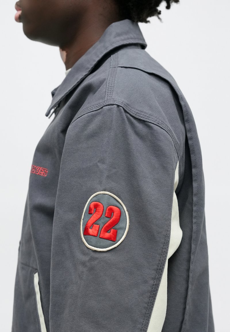 Gray work jacket featuring a prominent red "22" embroidered patch on the shoulder, with contrasting ivory details along the side seams.