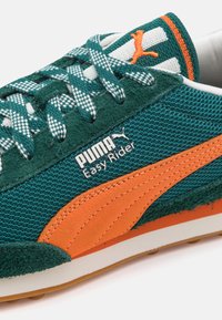Tenis verzi din plasă, cu accente de piele suede portocalie și o talpă intermediară albă. Logo-ul "PUMA Easy Rider" în alb pe lateral. Șireturi cu model vizibile.