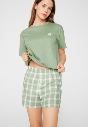 Jeune femme portant un t-shirt vert clair avec un petit motif de marguerite et un short à carreaux verts et blancs, soulevant légèrement son t-shirt.