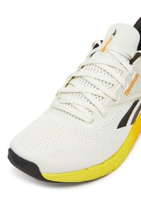 Balti bėgimo batai su tinklelio viršutine dalimi, juodais akcentais ir geltonu guminiu padu. Yra raišteliai, praeinantys per sustiprintas akių skylutes.