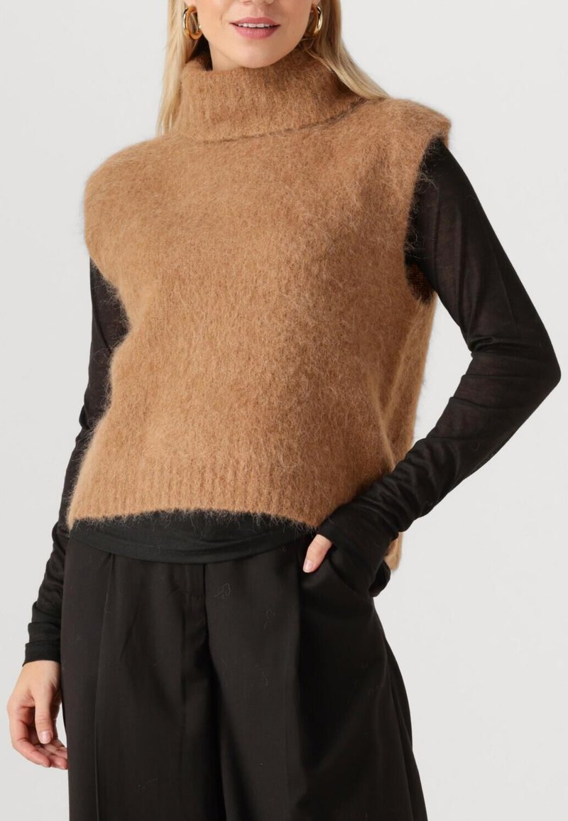 Frau trägt einen beige-flauschigen ärmellosen Rollkragenpullover über einem schwarzen Langarmshirt und schwarzen Hosen, eine Hand in der Tasche.