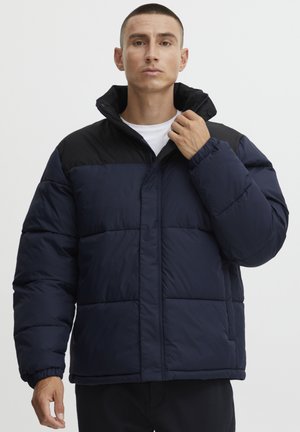 Navy puffer jacket with black upper panel, quilted pattern, high collar, and zip closure. Features elastic cuffs and a relaxed fit.