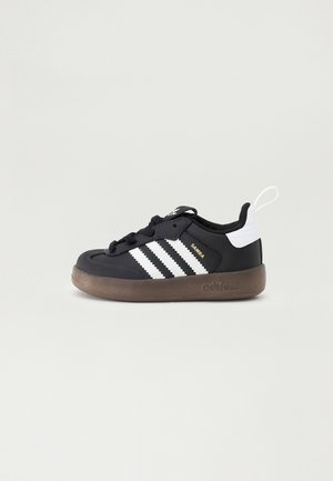 Sort Adidas Samba sneaker med hvide striber, brun sål, hvid hælflap og sorte snørebånd på ensartet lyst baggrund.