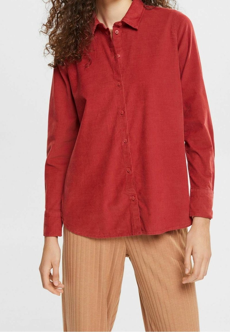 Chemise en velours côtelé rouge à boutons avec un col à revers et des manches longues. Présente une coupe décontractée et une texture lisse. Portée avec un pantalon beige côtelé.