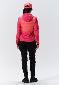 Lyserød jakke med quiltet ryg, der har en hætte, parret med sorte bukser og sneakers. Stoffet ser let og åndbart ud.