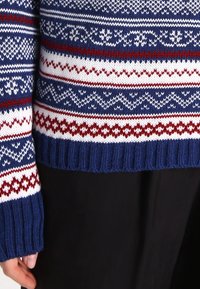 Sweter dzianinowy o niebieskiej bazie, zdobiony białymi i czerwonymi wzorami geometrycznymi. Ściągacze przy mankietach i hemie. Teksturowany materiał.