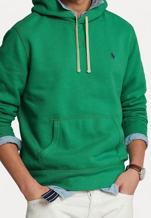Man draagt een groene hoodie over een lichtblauw spijkerblouse met opgerolde mouwen, witte broek en een bruine polshorloge, handen in de zakken.