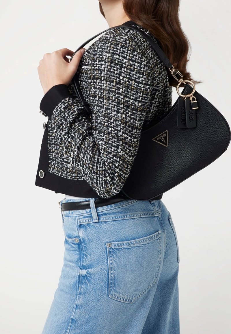 Femme portant une veste en tweed noire et blanche et un jean bleu clair tient un sac à main noir Guess sur son épaule.