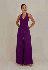 Purple halter jumpsuit with a fitted bodice and wide legs. Made of smooth fabric, features pleats at the waist, and paired with gold sandals.