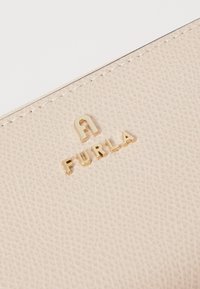 Beige Ledergeldbeutel mit strukturierter Oberfläche, ausgestattet mit einem goldenen, geprägten Logo "FURLA" in der Mitte. Gestichelte Kanten sind sichtbar.