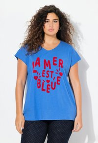 Modrá bavlněná tričko s červen�ým textem „L'AMER EST BLEU“ a srdíčky. Krátké rukávy, kulatý výstřih, volný střih. Nosené s vzorovanými tmavými kalhotami.