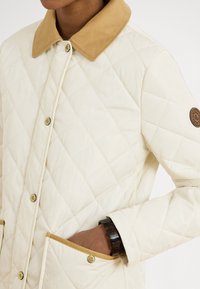 Jachetă albă matlasată cu guler din catifea dură bej, cu nasturi auriu și două buzunare laterale; țesătura are o textură netedă.