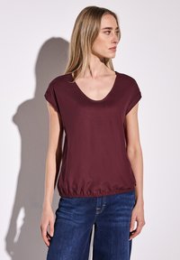 Top bordeaux à manches courtes avec un décolleté en V profond et une taille élastique, associé à un jean bleu à jambes larges, texture lisse et coupe décontractée.
