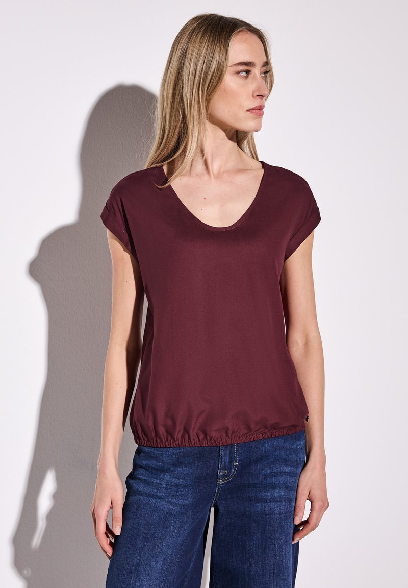 Top bordeaux à manches courtes avec un décolleté en V profond et une taille élastique, associé à un jean bleu à jambes larges, texture lisse et coupe décontractée.