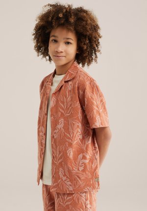 Fiatal, göndör hajú személy rövid ujjú narancssárga inget és hozzáillő, levelekkel mintázott rövidnadrágot visel fehér póló fölött.