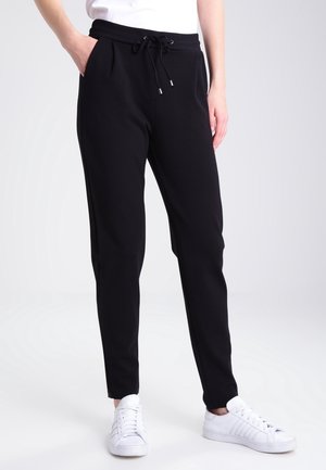 Person trägt schwarze Kordelzughose mit Taschen und weiße Turnschuhe, steht vor einem schlichten hellen Hintergrund.