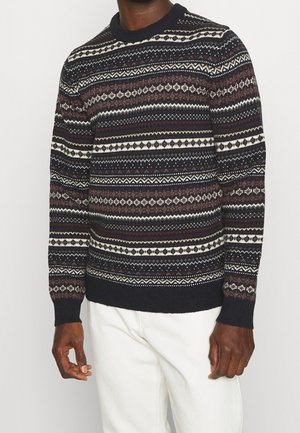 Mann trägt schwarzen Pullover mit Rundhalsausschnitt und horizontalen weißen, beigen und weinroten geometrischen Mustern sowie weiße Hose vor einfachem Hintergrund.