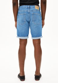 Shorts en denim bleu avec revers, design à cinq poches et une patch en cuir marron au dos de la taille. Texture lisse.