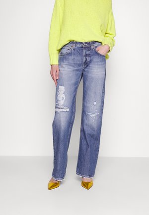 Person trägt hellblaue, ausgewaschene, weite Jeans, einen neon-gelben Strickpullover und spitze, metallisch goldene High Heels, steht vor weißem Hintergrund.