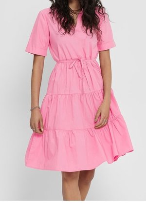 Kobieta ubrana w różową sukienkę do kolan z krótkimi rękawami, paskiem podkreślającym talię i warstwową falbanowaną spódnicą, stojąca na jednolitym tle.