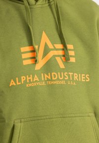 Sweat à capuche vert olive avec un logo graphique orange représentant un "A" stylisé et le texte "ALPHA INDUSTRIES" au-dessus d'une poche avant.
