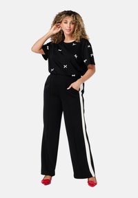 Zwarte top met witte kruisdessins, gecombineerd met zwarte wijde broek met witte zijkanten. Model draagt rode schoenen.