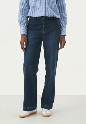 Denim jeans in dark blue with a straight-leg fit, featuring subtle stitching and a classic five-pocket design. Paired with white sneakers.