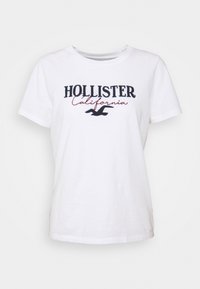 Bílý bavlněný tričko s nápisem "HOLLISTER California" v námořnické modři a vínové barvě, s černým logem ptáka, krátkými rukávy a kulatým výstřihem.