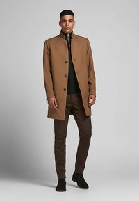 Brown wool blend coat with a tailored design, featuring a high collar, button closures, and worn over a black zip-up sweater and brown pants.
