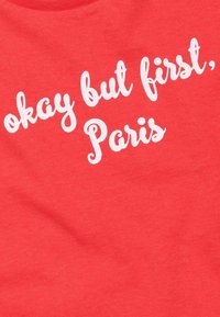 Rotes Baumwoll-T-Shirt mit weißem, geschwungenem Text, der "Okay, aber zuerst Paris" lautet. Glatte Textur mit rundem Ausschnitt.
