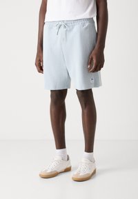 Shorts bleu clair côtelés avec une taille élastique et un cordon de serrage. Dotés d'un petit patch logo sur la jambe gauche. Portés avec des baskets blanches.