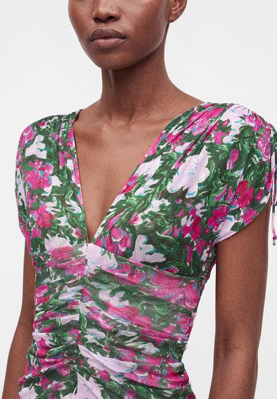Femme portant une robe fleurie froncée aux couleurs rose, verte et blanche, avec des manches courtes froncées et un décolleté en V profond.