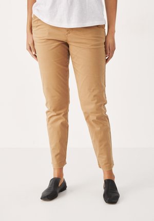 Unterkörper einer Person, die beige, schmal geschnittene Hose und schwarze Slip-On-Schuhe trägt, stehend vor einem schlichten hellen Hintergrund.