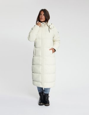 Vrouw draagt een lange witte donsjas met capuchon, blauwe spijkerbroek en zwarte veterlaarzen, staand met één hand in de zak.