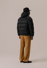 Doudoune noire avec capuche, en tissu texturé, et design matelassé horizontal, associée à un pantalon beige ample en tissu texturé et des chaussures noires.