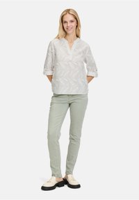 Blusa bianca con motivo a foglia, scollo a V e maniche arrotolate, abbinata a jeans skinny verde chiaro e scarpe crema con suole nere.