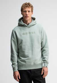 Jonge man met krullend haar, gekleed in een lichtgroene hoodie met de tekst "BUTCHER OF BLUE", staand tegen een effen witte achtergrond.