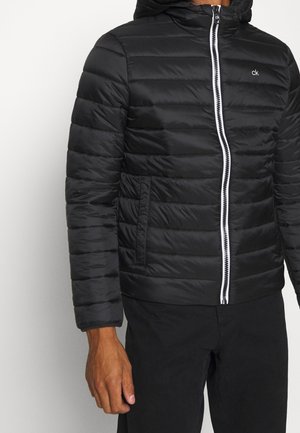 Veste matelassée noire avec fermeture éclair blanche et petit logo "ck", portée avec un pantalon noir, montrant le haut du torse et le bras gauche.