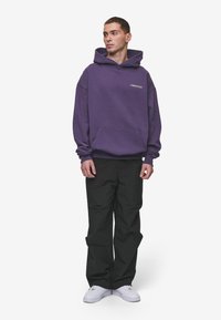 Sudadera morada con un bolsillo frontal y un logo blanco, combinada con pantalones negros de corte holgado y zapatillas blancas. Ajuste slim, textura suave.
