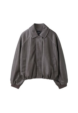 Jeune femme aux longs cheveux portant une veste texturée foncée, une mini-jupe assortie, un crop top noir, des bottes noires montant jusqu'aux genoux et des chaussettes grises.