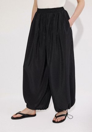 Γυναίκα που φοράει φαρδιά μαύρα χαρέμ παντελόνια με ελαστική μέση και μανσέτες, συνδυασμένα με μαύρα σαγιονάρες και λευκό τοπ, με τα χέρια στις τσέπες.