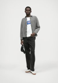 Camisa de ganga cinza sobre uma t-shirt branca com gráfico azul, emparelhada com jeans pretos e ténis brancos com solas de borracha. Asegurando uma cap preta.