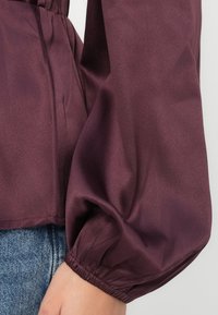 Burgundy, kiiltävä paita puffihihnoilla ja elastisilla hihansuilla, jossa on sileä rakenne ja khokkautettu vyötärö istuvan muodon saavuttamiseksi.