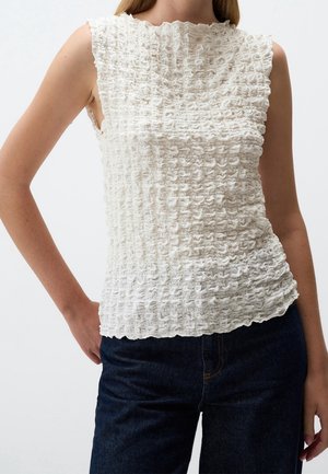 Woman wearing a sleeveless white textured top with ruffled edges and dark blue pants, standing with one hand on hip.