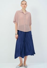 Camicia button-up rosa chiaro trasparente con maniche corte, abbinata a una gonna plissettata blu navy e scarpe ballerine argento.
