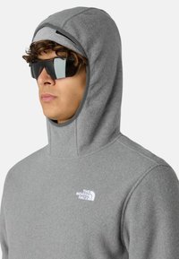 Szara polarowa bluza z wysokim kołnierzem i wbudowanym kapturem. Posiada logo w białym kolorze na piersi oraz gładką fakturę. Noszona w ciemnych okularach przeciwsłonecznych.