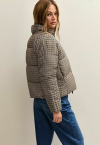 Bruine en beige geruite pufferjack met een hoge kraag, met een cropped ontwerp en gestructureerde stof, gedragen over blauwe denim jeans.