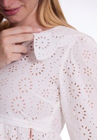Witte jurk met bloemen uitsnijdingen, gegolfde kraag en korte pofmouwen. De stof lijkt licht en textuur te hebben. Zijdelingse ritssluiting zichtbaar.